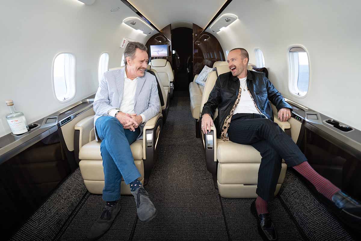 Bryan Cranston and Aaron Paul laughing on Boeing jet, next to Dos Hombres bottle 
