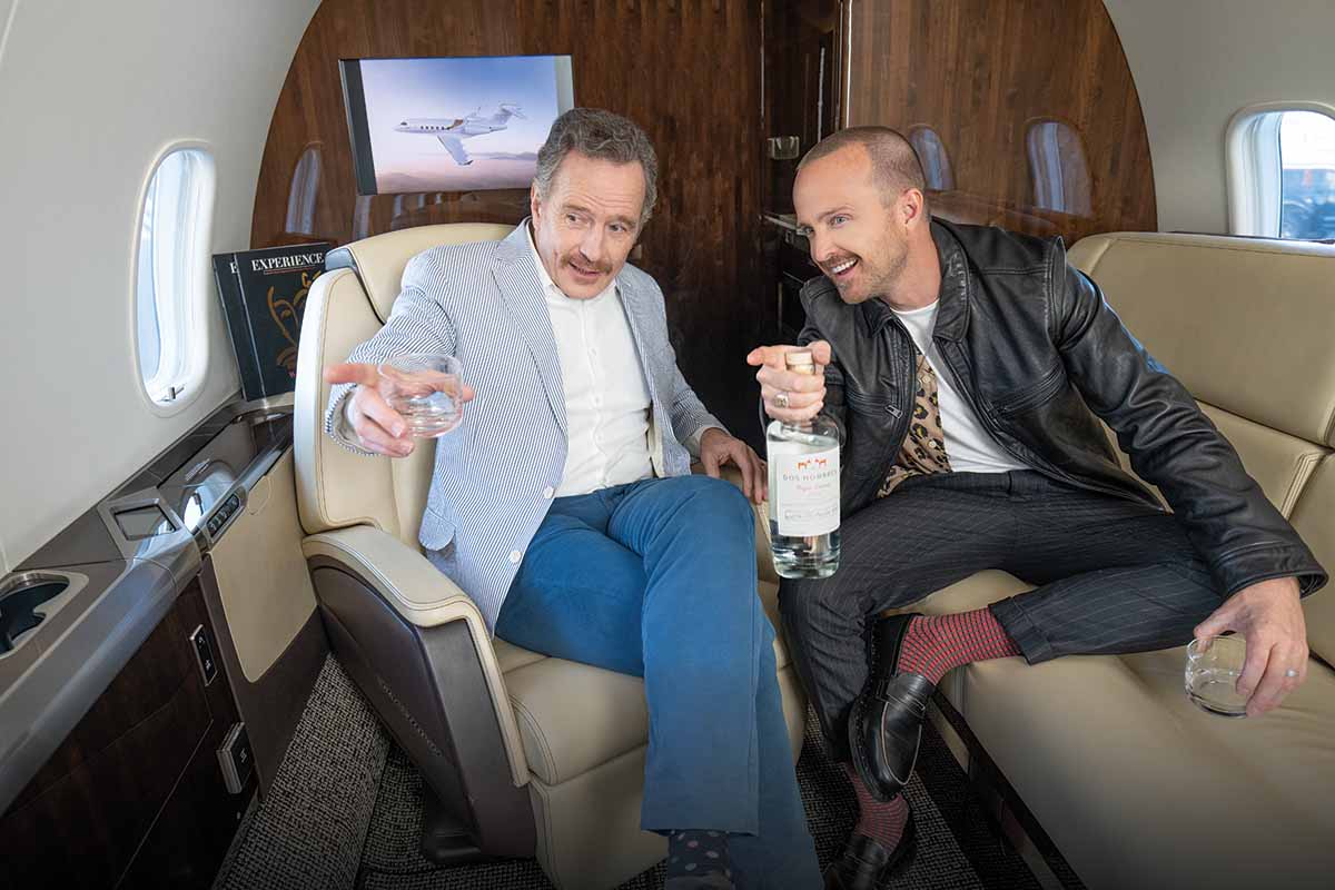 Bryan Cranston and Aaron Paul pointing at something offscreen. Aaron is holding a Dos Hombres bottle.