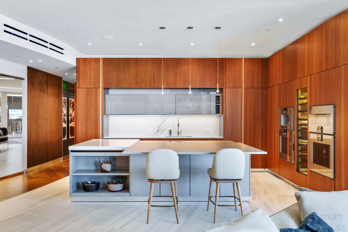 Picture of a Bentley Residences kitchen. There are two chairs at the white-marble counter. The walls are cerused wood, and the appliances slot neatly inside of them. 