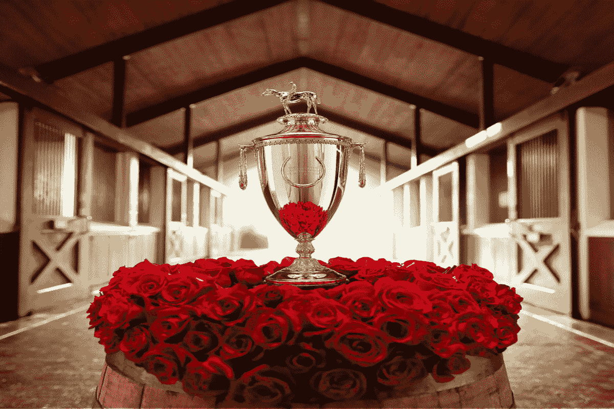 Kentucky Derby gold trophy on a pile of roses, elevated by a barrel. In the background, horse stables. 